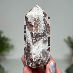 May include: A polished, rectangular crystal tower with a pointed top. The stone features a mix of white, brown, and reddish-brown patterns. The crystal is held in a person's hand, showcasing its natural beauty and unique veining.