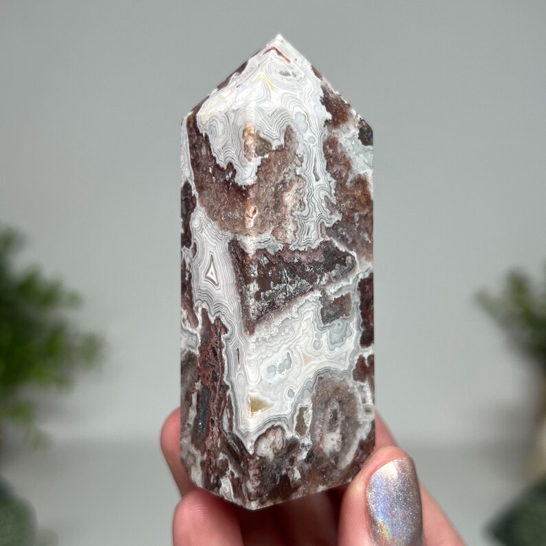 May include: A polished, rectangular crystal tower with a pointed top. The stone features a mix of white, brown, and reddish-brown patterns. The crystal is held in a person's hand, showcasing its natural beauty and unique veining.