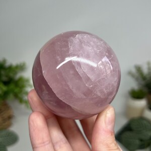 May include: A polished rose quartz sphere, showcasing a soft pink hue with translucent areas. The sphere is held in a hand, with a blurred background of greenery. This crystal ball is a decorative item.