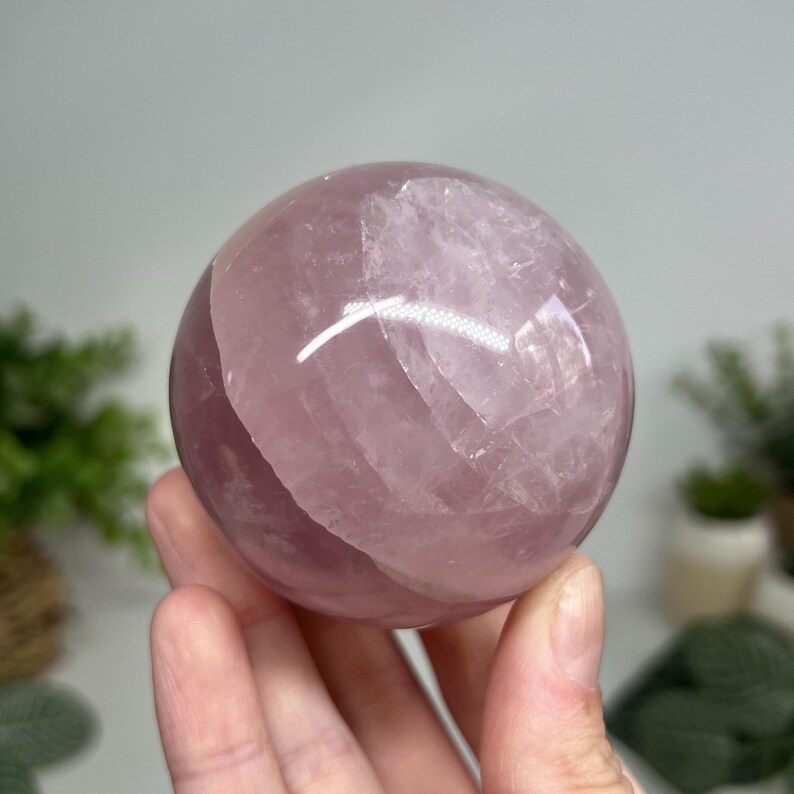 May include: A polished rose quartz sphere, showcasing a soft pink hue with translucent areas. The sphere is held in a hand, with a blurred background of greenery. This crystal ball is a decorative item.