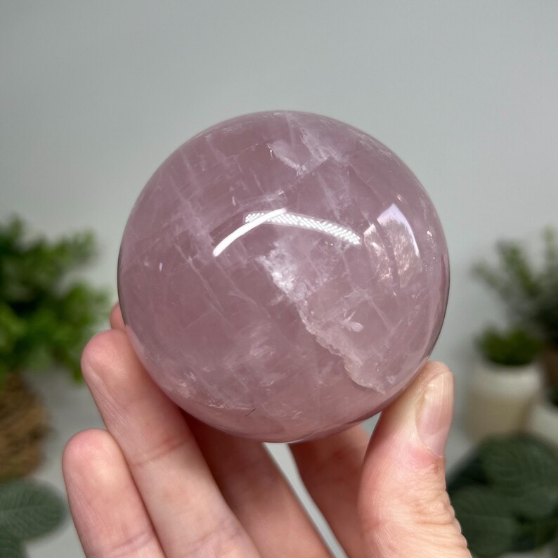 May include: A polished, translucent rose quartz sphere. The sphere is a soft pink color with natural inclusions and a smooth, reflective surface. The crystal ball is held in a hand.