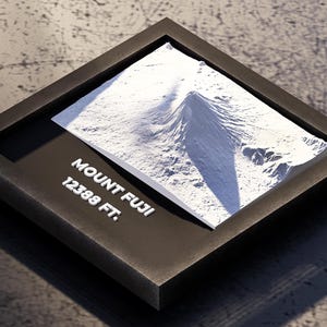 May include: A dark brown framed relief model of Mount Fuji, with the text "MOUNT FUJI 12388 FT." in white. The model depicts the mountain's terrain in white, set against a dark background.