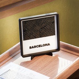 May include: A framed, black and white relief map of Barcelona, Spain, with the city's name in bold letters. The map is set against a white background and displayed on a wooden surface.