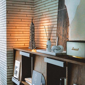 May include: A black and white Empire State Building model sits on a wooden shelf, next to a white marble tray with a decorative knot and a diffuser. A Marshall speaker is on the right. The background is a brick wall.