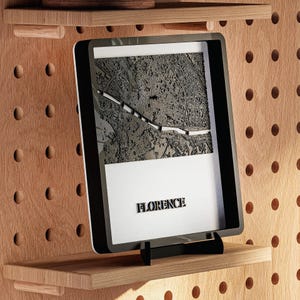 May include: A framed, three-dimensional map of Florence, Italy, in black and white, displayed on a wooden shelf. The word "FLORENCE" is printed in bold, black letters at the bottom of the frame. The frame is black with a white border.