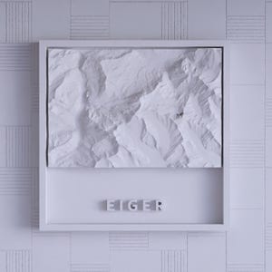 Puede incluir: Un mapa en relieve blanco enmarcado de una cordillera, posiblemente el Eiger, con texturas y sombras detalladas. La palabra "EIGER" se muestra en letras de bloque debajo del mapa, sobre un fondo blanco.