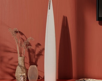 May include: A white wooden model of a skyscraper with a tall, slender design. The model is sitting on a table with a vase of dried flowers, a wicker basket, and a book.