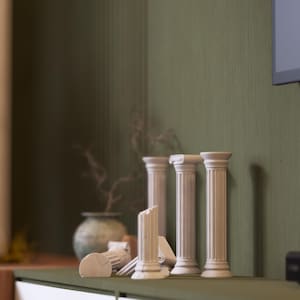 Puede incluir: Una colección de pequeñas columnas blancas de estilo clásico, algunas enteras y otras rotas, dispuestas sobre una estantería verde. Un pequeño jarrón redondo verde y ramas secas en el fondo. El conjunto se encuentra contra una pared verde texturizada.