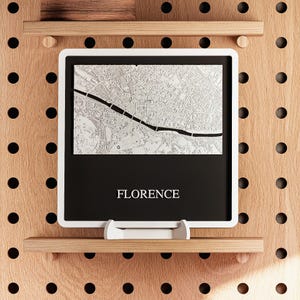 May include: A framed map of Florence, Italy, with a white border. The map is in black and white, showing the city's layout. The word "FLORENCE" is printed in white at the bottom of the black section. The frame is displayed on a wooden pegboard.