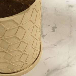 Op de afbeelding: Een beige plantenpot met een geometrisch patroon. De pot is gemaakt van een getextureerd materiaal en heeft een licht afgeronde bodem.