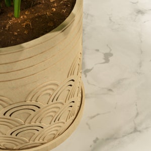May include: A beige, cylindrical planter with a decorative wave pattern carved into the lower portion. The planter is filled with soil and has green plant stems visible. The design is reminiscent of Japanese art.