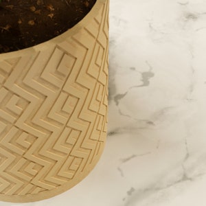 May include: A beige, geometric patterned planter with soil visible at the top. The planter has a repeating square and zig-zag design. The background is a white marble-like surface.