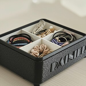 May include: A black, textured organizer box with four compartments, filled with hair accessories. The box contains scrunchies, hair ties, and barrettes in various colors. The word "PANIER" is embossed on the side.