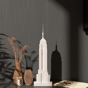 May include: A white Empire State Building model stands tall, next to a woven vase with dried plants, a decorative openwork vase, and a small candle holder. The objects are arranged on a dark surface against a dark grey wall.