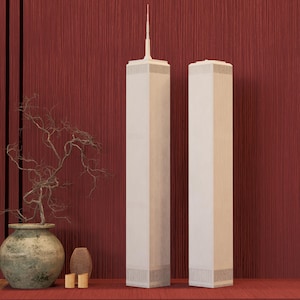 Könnte beinhalten: Zwei cremefarbene, vertikale Skulpturen der World Trade Center Türme stehen auf einer roten Oberfläche vor einem roten, strukturierten Hintergrund. Im Vordergrund befinden sich eine dekorative Vase mit einer Pflanze und andere dekorative Gegenstände.