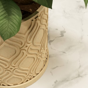 May include: A beige, patterned plant pot with a curved design. The pot has a circular base and contains soil and green plant leaves. The background is a white marble surface.