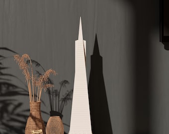 Op de afbeelding: Een wit model van het Transamerica Pyramid-gebouw, een rieten vaas met gedroogde takken, een rieten mand en een boek op een donkergrijs oppervlak.