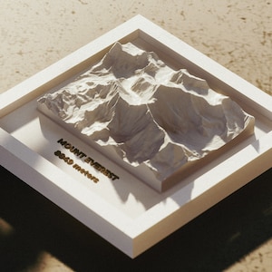 Puede incluir: Un modelo de yeso blanco del Monte Everest en un marco blanco. El modelo está elevado del marco y tiene una inscripción dorada que dice "MOUNT EVEREST 8848 METROS".