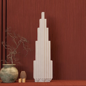 May include: A cream-coloured, Art Deco-style sculpture resembling a skyscraper stands on a red surface against a textured red wall. A decorative vase with a plant and other decorative items are to the left.