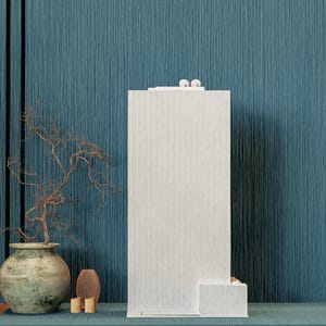 Könnte beinhalten: Ein hoher, cremefarbener, strukturierter Schrank mit einem kleinen passenden Sockel. Der Schrank hat ein minimalistisches Design mit einer dekorativen Oberseite. Im Hintergrund befindet sich eine blaue, strukturierte Wand sowie eine dekorative Vase und ein kleiner Ast.