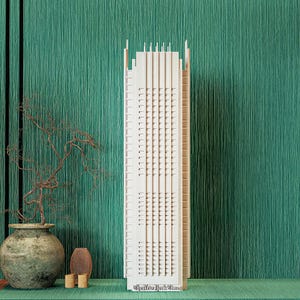 Könnte beinhalten: Ein detailliertes, cremefarbenes Modell eines Wolkenkratzers mit zahlreichen Fenstern und architektonischen Details. Es steht vor einem türkisfarbenen, strukturierten Hintergrund, mit einer dekorativen Vase und Zweigen. Der Text "The New York Times" ist sichtbar.