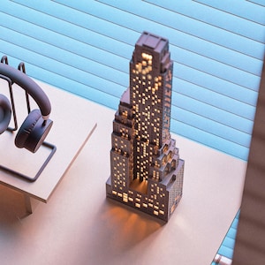 May include: A miniature brown skyscraper model with illuminated windows, showcasing a detailed architectural design. The model is placed on a light-colored surface, with a pair of headphones in the background. The building's windows glow with a warm light.