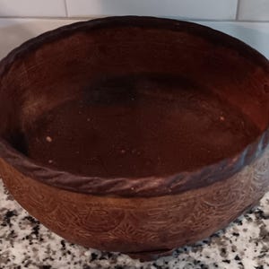 Könnte beinhalten: Eine runde, braune Metallschale mit zwei verzierten Griffen. Die Schale hat ein strukturiertes, geprägtes Muster um den Boden und ein gedrehtes Seildesign um den Rand. Der Innenraum ist dunkelbraun.