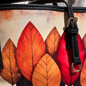 K&ouml;nnte beinhalten: Tragetasche mit einem lebendigen Herbstblatt-Design. Die Tasche zeigt gro&szlig;e, stilisierte Bl&auml;tter in Rot-, Orange- und Braunt&ouml;nen vor einem cremefarbenen Hintergrund. Sie hat schwarze Griffe mit goldfarbener Hardware.