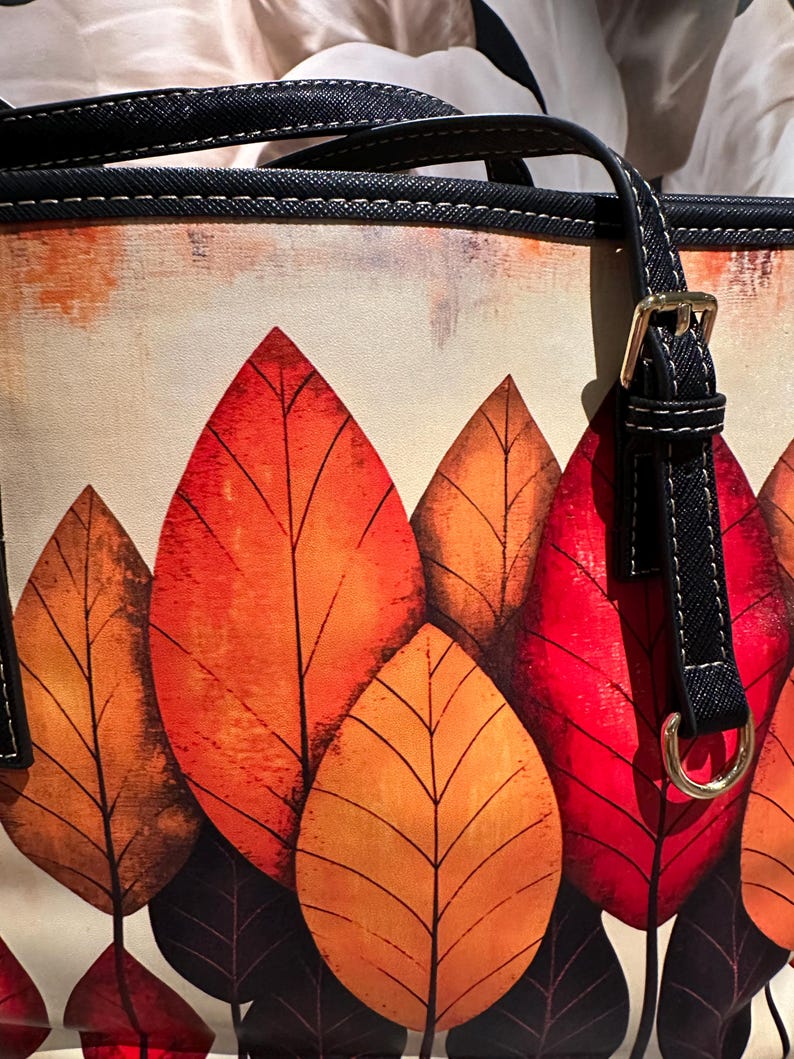 K&ouml;nnte beinhalten: Tragetasche mit einem lebendigen Herbstblatt-Design. Die Tasche zeigt gro&szlig;e, stilisierte Bl&auml;tter in Rot-, Orange- und Braunt&ouml;nen vor einem cremefarbenen Hintergrund. Sie hat schwarze Griffe mit goldfarbener Hardware.