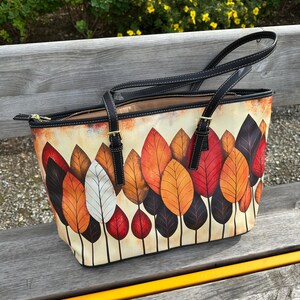 K&ouml;nnte beinhalten: Tragetasche mit einem farbenfrohen Herbstblatt-Design. Die Tasche hat einen cremefarbenen Hintergrund mit einem Muster aus roten, orangefarbenen, braunen und wei&szlig;en Bl&auml;ttern. Sie hat schwarze Griffe und Verzierungen sowie einen Rei&szlig;verschluss.