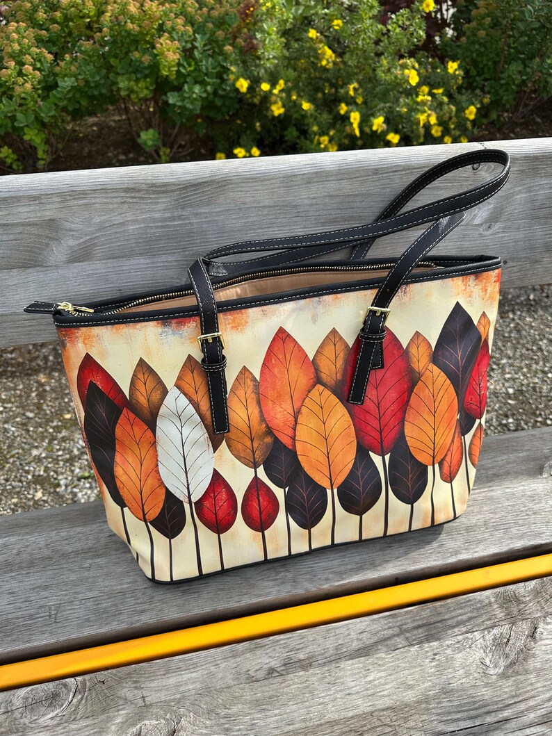 K&ouml;nnte beinhalten: Tragetasche mit einem farbenfrohen Herbstblatt-Design. Die Tasche hat einen cremefarbenen Hintergrund mit einem Muster aus roten, orangefarbenen, braunen und wei&szlig;en Bl&auml;ttern. Sie hat schwarze Griffe und Verzierungen sowie einen Rei&szlig;verschluss.