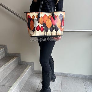 K&ouml;nnte beinhalten: Eine Tragetasche mit einem farbenfrohen Blattdesign. Die Tasche hat einen cremefarbenen Hintergrund mit einem Muster aus stilisierten Bl&auml;ttern in Rot-, Orange- und Braunt&ouml;nen. Die Tasche hat schwarze Griffe und Verzierungen. Der Text "Heartfelt Whisper" ist auf der Unterseite der Tasche aufgedruckt.