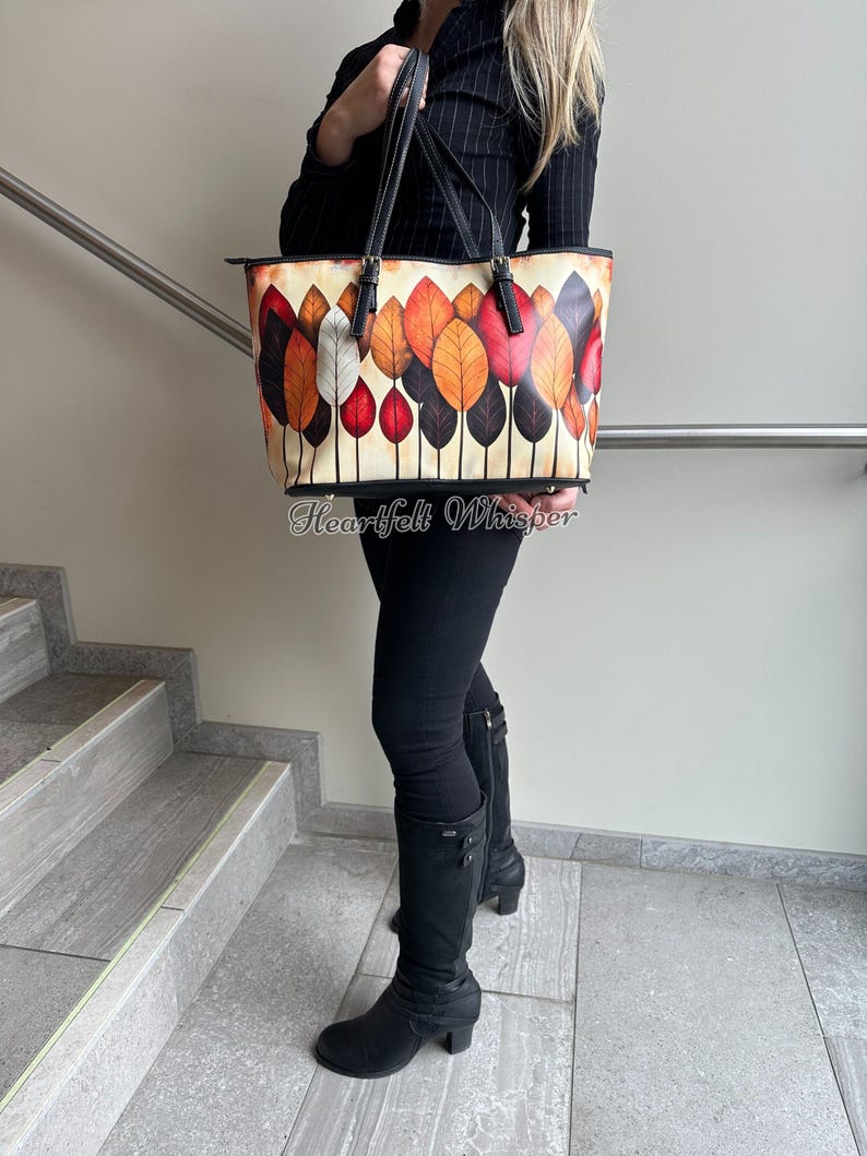 K&ouml;nnte beinhalten: Eine Tragetasche mit einem farbenfrohen Blattdesign. Die Tasche hat einen cremefarbenen Hintergrund mit einem Muster aus stilisierten Bl&auml;ttern in Rot-, Orange- und Braunt&ouml;nen. Die Tasche hat schwarze Griffe und Verzierungen. Der Text "Heartfelt Whisper" ist auf der Unterseite der Tasche aufgedruckt.
