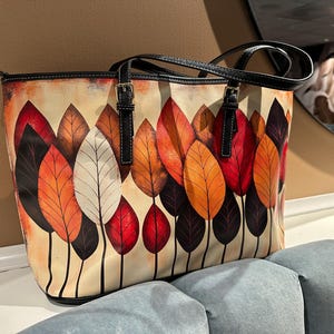K&ouml;nnte beinhalten: Tragetasche mit farbenfrohem Blattdesign. Die Tasche hat einen cremefarbenen Hintergrund mit einem Muster aus stilisierten Bl&auml;ttern in Rot-, Orange- und Braunt&ouml;nen. Sie hat schwarze Griffe und Verzierungen mit silberfarbenen Schnallen.