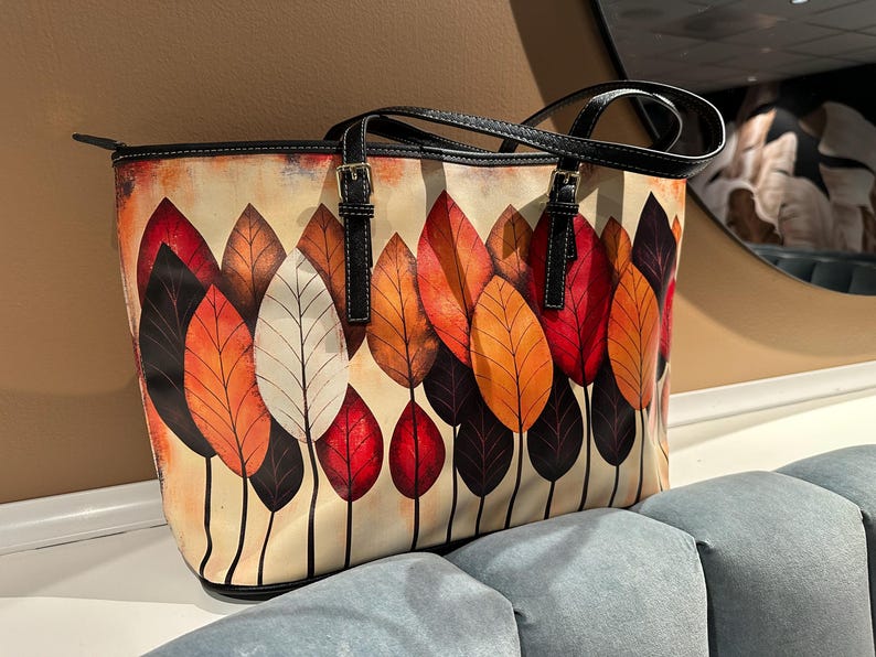 K&ouml;nnte beinhalten: Tragetasche mit farbenfrohem Blattdesign. Die Tasche hat einen cremefarbenen Hintergrund mit einem Muster aus stilisierten Bl&auml;ttern in Rot-, Orange- und Braunt&ouml;nen. Sie hat schwarze Griffe und Verzierungen mit silberfarbenen Schnallen.