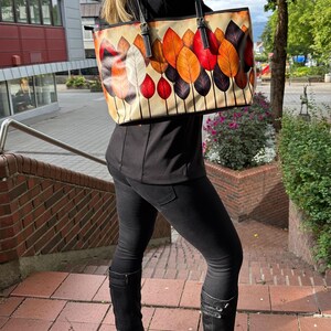 K&ouml;nnte beinhalten: Beige Tragetasche mit einem farbenfrohen Blattdesign in Orange-, Rot- und Lilat&ouml;nen. Die Tasche hat schwarze Griffe und eine rechteckige Form. Die Tasche wird &uuml;ber der Schulter getragen.