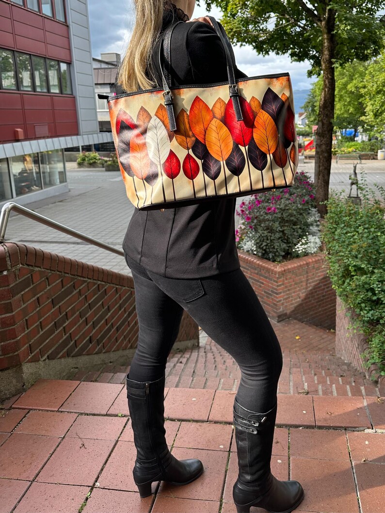 K&ouml;nnte beinhalten: Beige Tragetasche mit einem farbenfrohen Blattdesign in Orange-, Rot- und Lilat&ouml;nen. Die Tasche hat schwarze Griffe und eine rechteckige Form. Die Tasche wird &uuml;ber der Schulter getragen.