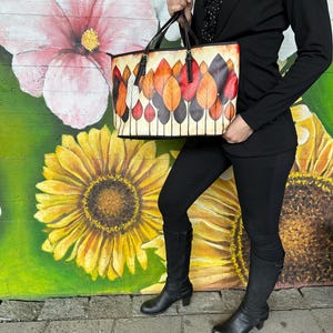 K&ouml;nnte beinhalten: Eine rechteckige Tragetasche mit einem farbenfrohen Blattdesign. Die Tasche hat einen cremefarbenen Hintergrund mit einem Muster aus stilisierten Bl&auml;ttern in Rot-, Orange- und Braunt&ouml;nen. Die Tasche hat schwarze Griffe und Verzierungen.