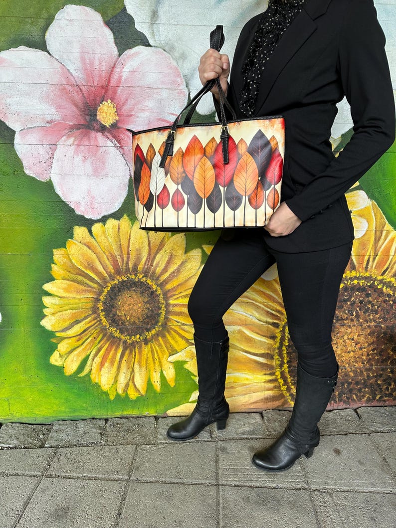 K&ouml;nnte beinhalten: Eine rechteckige Tragetasche mit einem farbenfrohen Blattdesign. Die Tasche hat einen cremefarbenen Hintergrund mit einem Muster aus stilisierten Bl&auml;ttern in Rot-, Orange- und Braunt&ouml;nen. Die Tasche hat schwarze Griffe und Verzierungen.