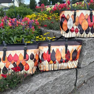 K&ouml;nnte beinhalten: Drei Tragetaschen mit einem Herbstblatt-Design. Die Taschen haben einen cremefarbenen Hintergrund mit bunten Bl&auml;ttern in Rot-, Orange- und Braunt&ouml;nen. Die Taschen haben schwarze Griffe und Riemen mit goldenen Schnallen.
