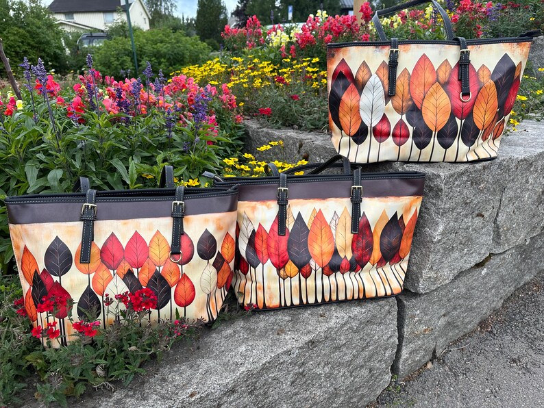 K&ouml;nnte beinhalten: Drei Tragetaschen mit einem Herbstblatt-Design. Die Taschen haben einen cremefarbenen Hintergrund mit bunten Bl&auml;ttern in Rot-, Orange- und Braunt&ouml;nen. Die Taschen haben schwarze Griffe und Riemen mit goldenen Schnallen.