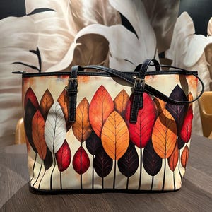 K&ouml;nnte beinhalten: Eine Tragetasche mit einem farbenfrohen Blattdesign. Die Tasche hat einen cremefarbenen Hintergrund mit einem Muster aus roten, orangefarbenen und lila Bl&auml;ttern. Sie hat schwarze Griffe und Riemen sowie einen Rei&szlig;verschluss. Die Tasche steht auf einem Holztisch.