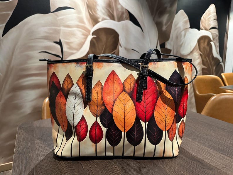 K&ouml;nnte beinhalten: Eine Tragetasche mit einem farbenfrohen Blattdesign. Die Tasche hat einen cremefarbenen Hintergrund mit einem Muster aus roten, orangefarbenen und lila Bl&auml;ttern. Sie hat schwarze Griffe und Riemen sowie einen Rei&szlig;verschluss. Die Tasche steht auf einem Holztisch.
