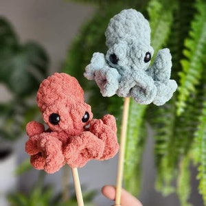 Puede incluir: Dos peluches de pulpo de crochet en palos de madera. Un pulpo es azul claro y el otro es coral.