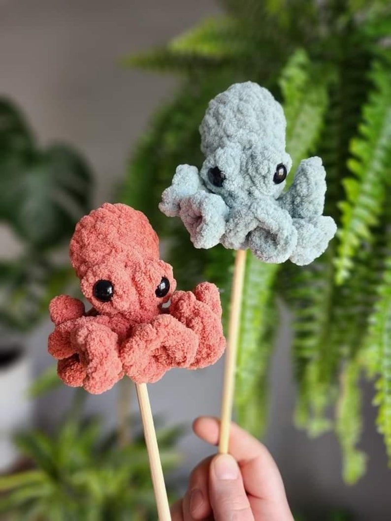 Puede incluir: Dos peluches de pulpo de crochet en palos de madera. Un pulpo es azul claro y el otro es coral.