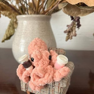 Puede incluir: Un pulpo de crochet rosa sentado en un sill&oacute;n de patrones marrones y beige, sosteniendo dos tazas peque&ntilde;as con tapas.