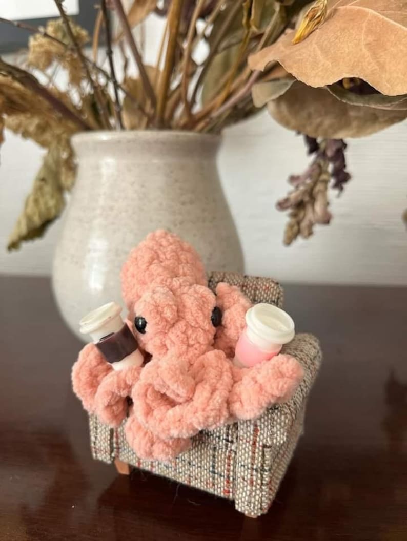 Puede incluir: Un pulpo de crochet rosa sentado en un sill&oacute;n de patrones marrones y beige, sosteniendo dos tazas peque&ntilde;as con tapas.