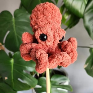 Puede incluir: Un juguete de pulpo de crochet de color rosa coral con ojos negros. El pulpo est&aacute; sentado en un palo de madera.