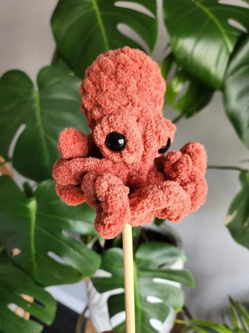 Puede incluir: Un juguete de pulpo de crochet de color rosa coral con ojos negros. El pulpo est&aacute; sentado en un palo de madera.