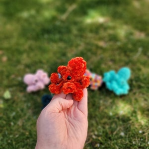 Puede incluir: Un peque&ntilde;o pulpo de crochet hecho de hilo rojo con ojos negros. El pulpo est&aacute; siendo sostenido en la mano de una persona.